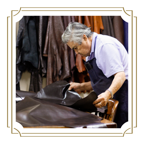 An older person with gray hair, wearing an apron, inspects or works with a piece of dark leather cloth.