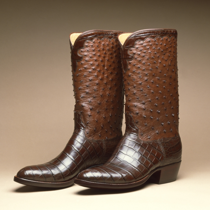 A pair of custom handmade cowboy boots with intricate leather detailing, including a textured shaft and smooth toe and heel, against a neutral background.