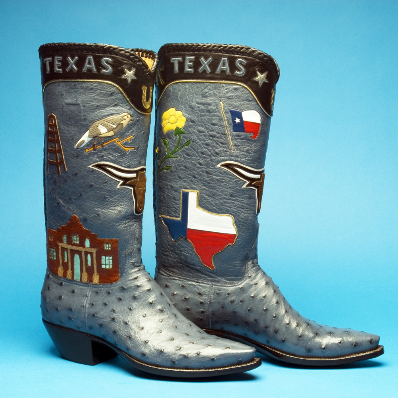 A pair of custom handmade cowboy boots embellished with Texas symbols such as the state flower, flag, map, a bull, oil rig, and Alamo on a blue background.