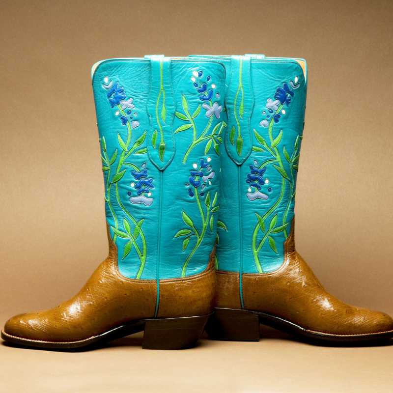 A stunning pair of handmade cowboy boots in blue and brown, featuring exquisite floral embroidery, displayed against a beige background.