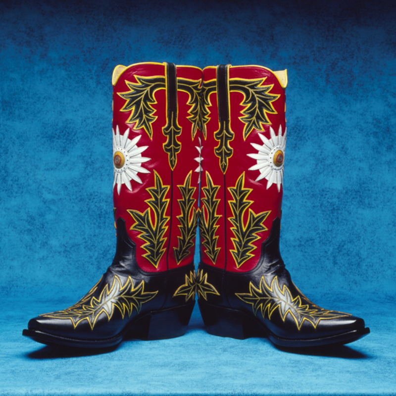 A pair of black, red, and yellow custom handmade cowboy boots with intricate floral and leaf designs against a blue background.