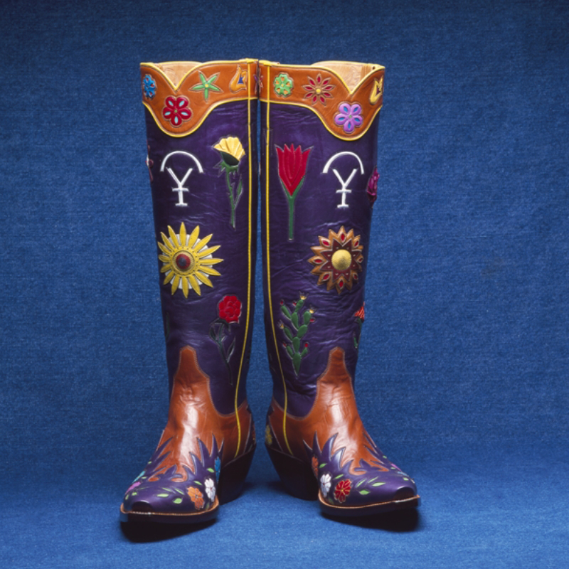 A pair of colorful, custom handmade cowboy boots with intricate floral designs and symbols, set against a plain blue background.