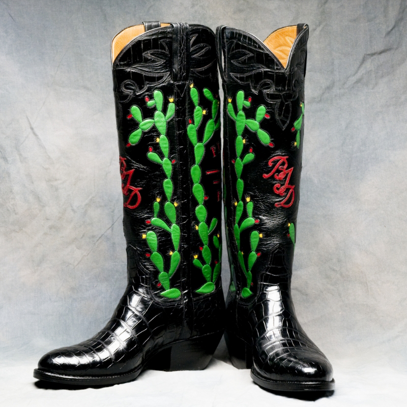 Custom handmade cowboy boots featuring green cactus designs and red initials stand out against a gray background, perfect for Western fashion enthusiasts.