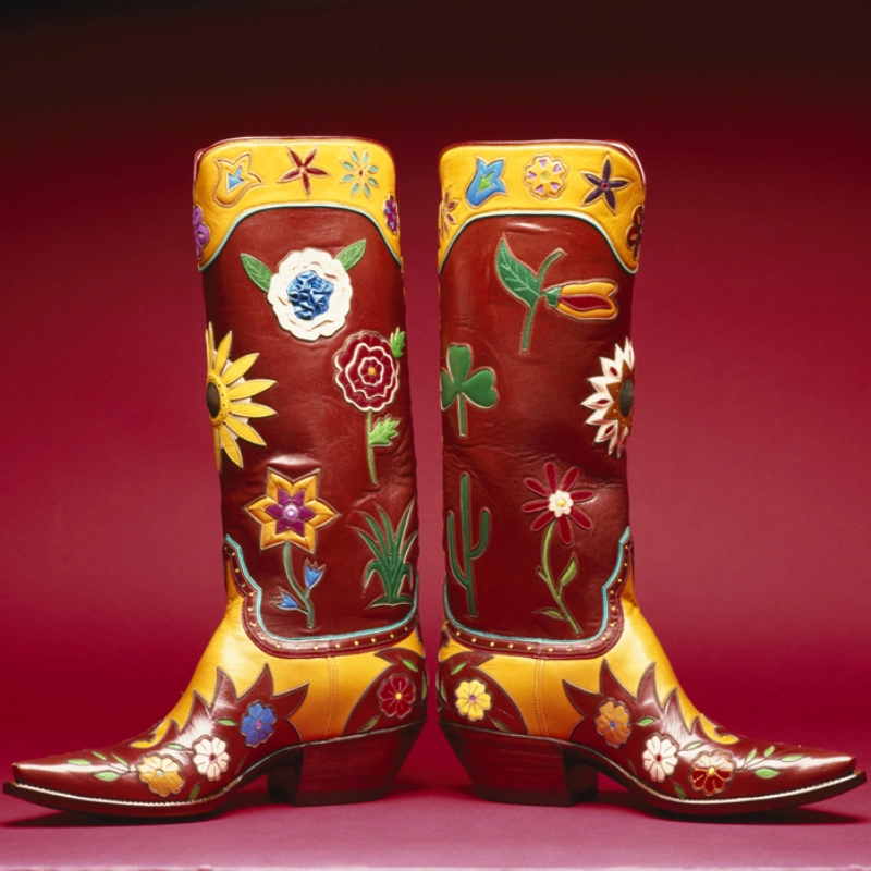 A pair of handmade cowboy boots in red and yellow, featuring colorful floral and plant designs against a maroon background.