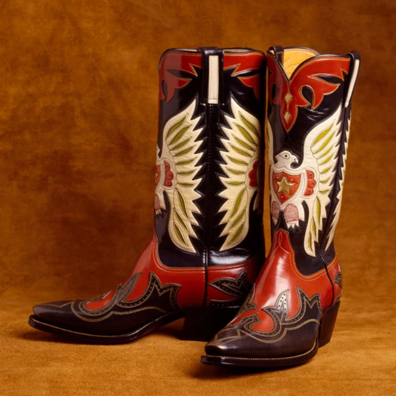 A pair of black and red handmade cowboy boots with intricate winged eagle designs on the sides, displayed against a brown background.