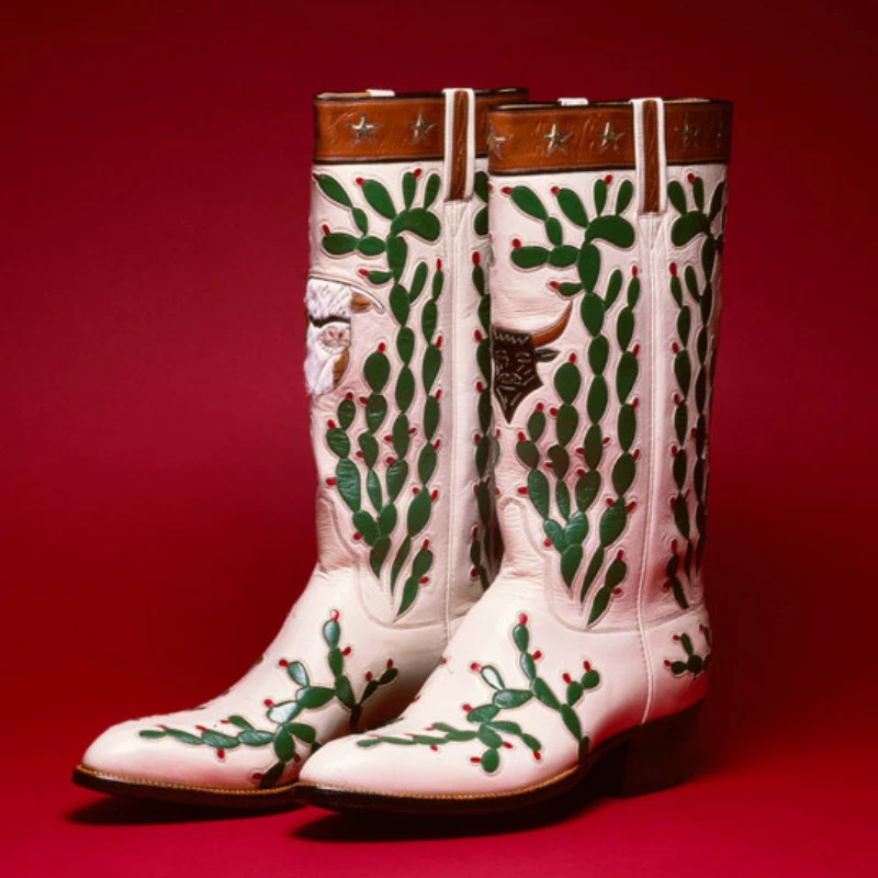 A pair of custom handmade cowboy boots in white, adorned with green cactus and red flower designs, set against a striking red background.
