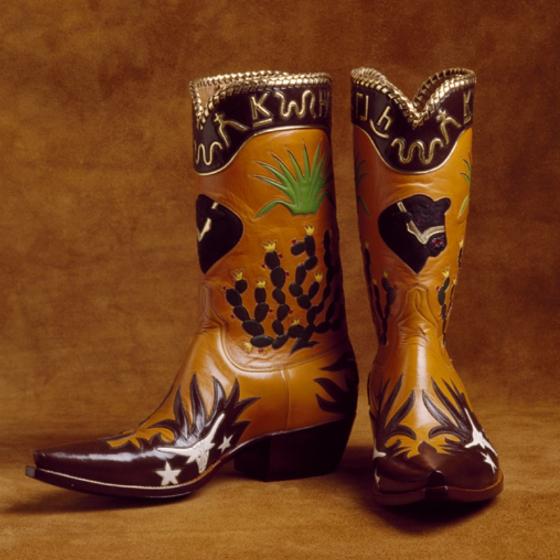 A pair of intricately designed handmade cowboy boots with cactus, steer, and star motifs on a brown background.