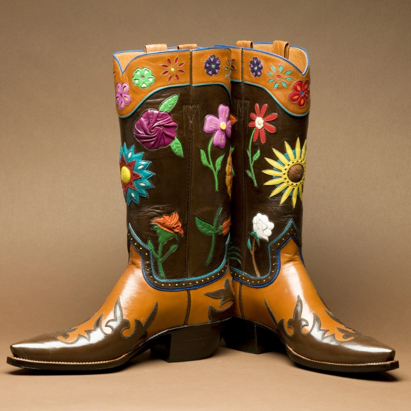A pair of brown handmade cowboy boots with colorful floral embroidery on the shafts and decorative stitching on the toes, crafted by skilled custom cowboy boot makers in Texas, set against a neutral background.