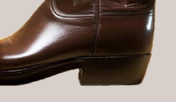 A close-up of the heel and lower portion of a smooth, shiny, brown leather boot against a beige background showcases the exquisite craftsmanship of handmade cowboy boots.